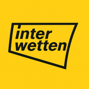 Persona sosteniendo un teléfono móvil con el logotipo amarillo de Interwetten en pantalla, con una tabla de cuotas deportivas desenfocada en el fondo, representando el uso de la plataforma de casino Interwetten.